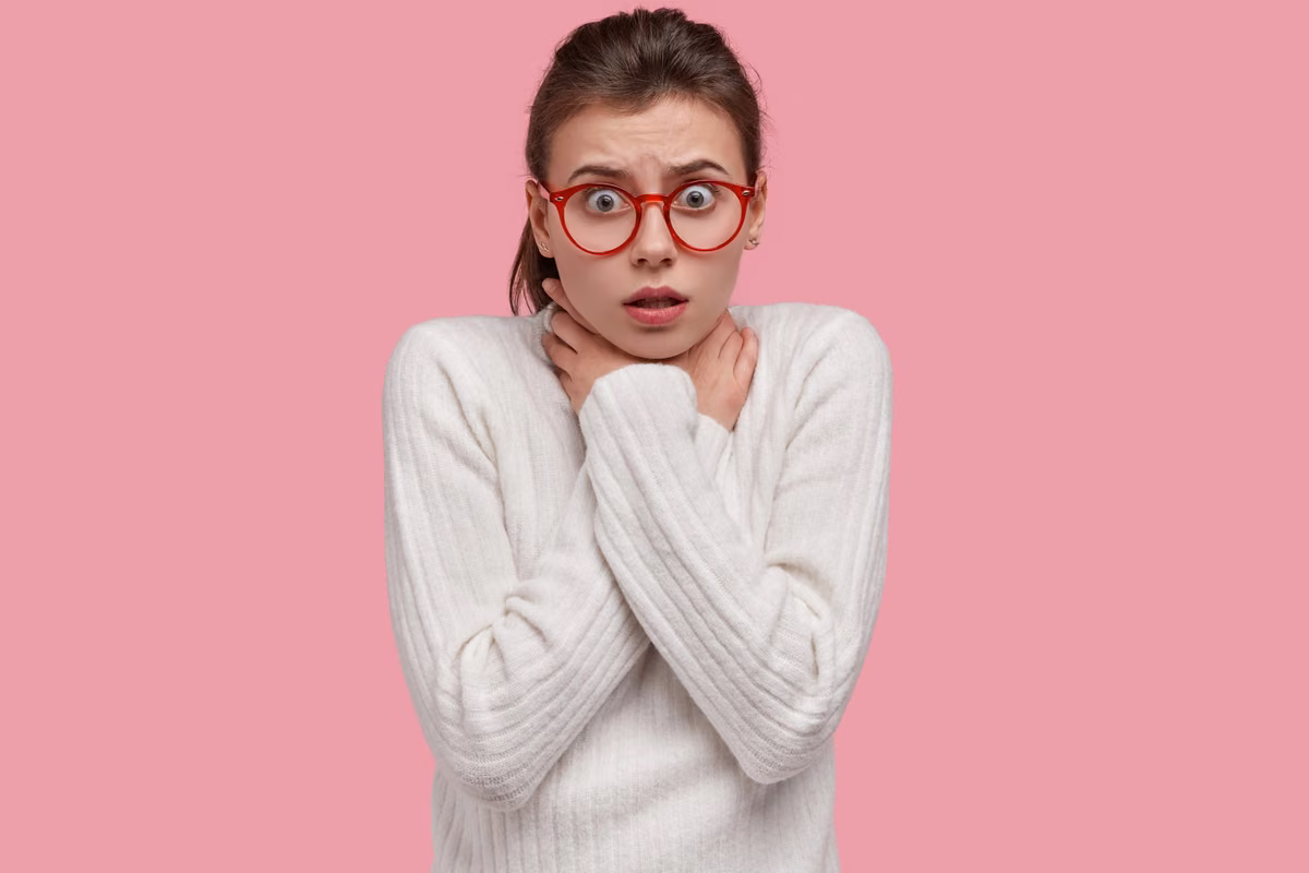 Woman in red glasses and a white jumper looking startled against a pink backdrop